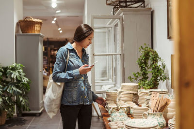 Young woman with mobile phone choosing crockery arranged on table in boutique