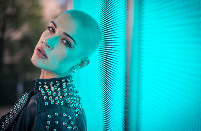 Portrait of young woman with shaved head standing against blue wall