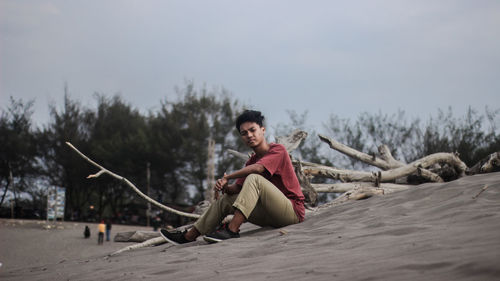Portrait of young man sitting on sand against sky
