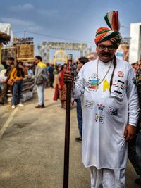 Portrait of man standing holding walking cane in traditional clothing against sky