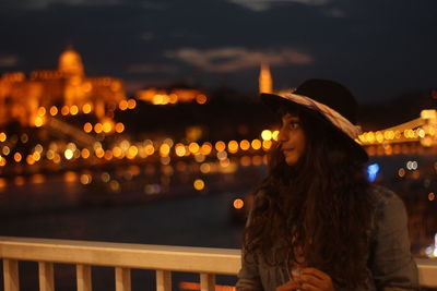 Young woman looking away while standing by railing with illuminated city in background at night