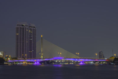 Illuminated bridge over river by buildings against sky at night