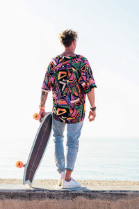 Back view of young hipster male skater in trendy summer outfit carrying skateboard while standing on embankment against sea and cloudless sky in summer day