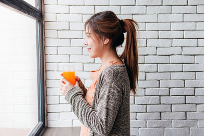 Side view of young woman holding drink glass by wall