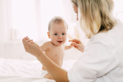 A cute baby is sitting on the bed with her arms outstretched and holding her mother's hands. 