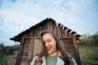 Beautiful girl with long hair in a grey trench coat next to an old wooden house outdoors in spring