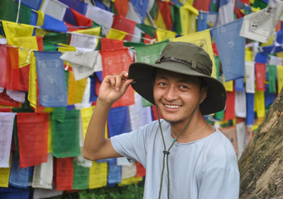 A happy asian young man looking sideways with touching hat, posing against buddhist prayer flags