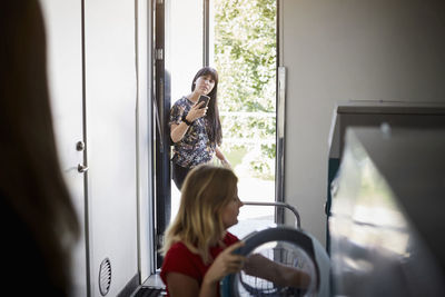 Woman using mobile phone at doorway and friend doing laundry