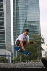 Young man doing stunt with skateboard against building in city