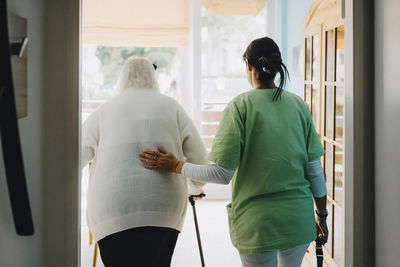Rear view of female nurse supporting elderly woman walking at retirement home
