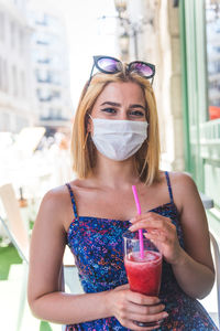 Portrait of young woman wearing mask drinking juice sitting at cafe