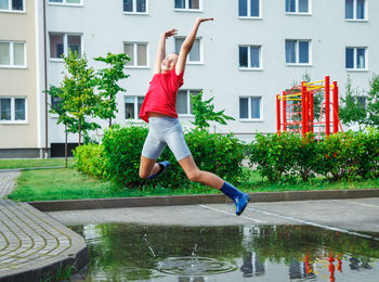 Cheerful girl jumping over puddle against building