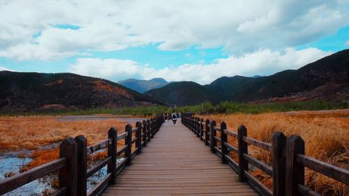 View of wooden footbridge leading towards mountains against sky