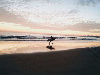 Man carrying surfboard while walking at beach during sunset