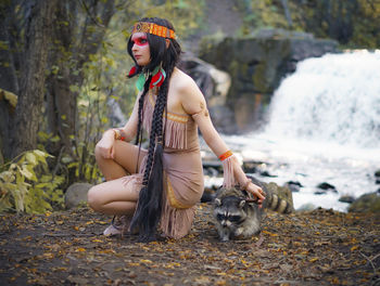 Young woman in traditional clothing looking away while crouching by raccoon in forest