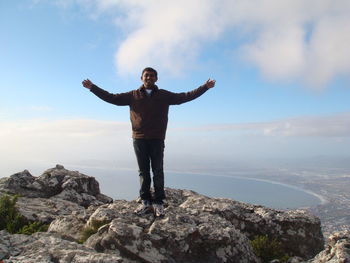 Full length of man with arms outstretched standing on rock against sky