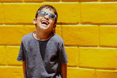 Cheerful boy wearing sunglasses standing against yellow brick wall on sunny day
