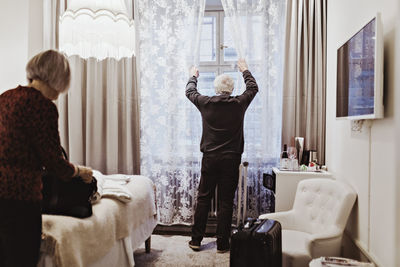 Senior man opening curtains while woman holding bag in hotel room