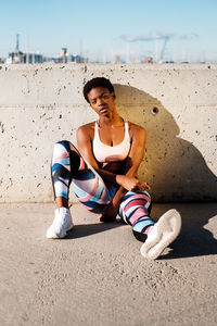 African american female athlete in flowered sport clothes and white sneakers looking at camera while sitting alone on concrete wall in sunbeams and resting after training in city