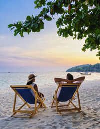 Rear view of woman sitting on lounge chair at beach against sky