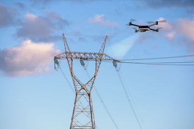 Low angle view of drone flying by electricity pylon against sky