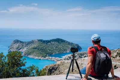 Rear view of man sitting by camera on retaining wall while looking at sea against sky