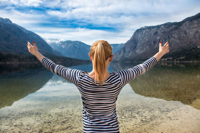 Rear view of woman standing with arms outstretched by lake against mountains