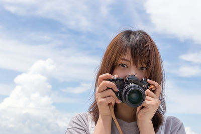 Portrait of young woman holding camera against cloudy sky