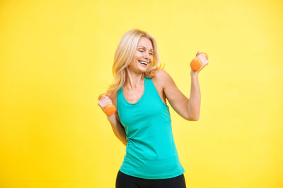 Young woman lifting dumbbell against yellow background