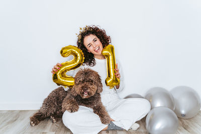 Portrait of smiling woman holding balloons sitting with dog against wall at home