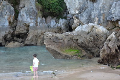 Rear view of boy standing on shore at beach against rock formations