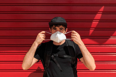 Unrecognizable trendy guy in black apparel putting on respiratory mask while looking at camera near colorful wall during coronavirus pandemic