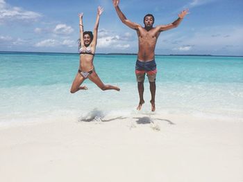 Full length of happy friends with arms raised jumping at beach against sky