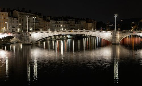 Bridge over river by illuminated buildings against sky at night