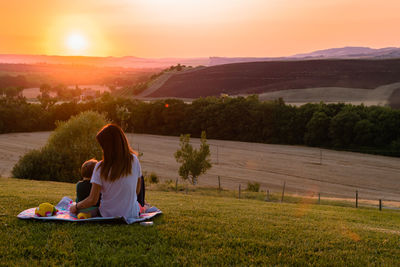 Mother and daughter sitting on field against sky during sunset