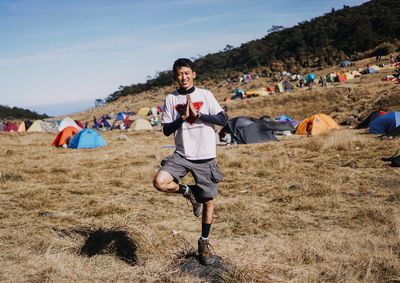 Portrait of young man exercising on grassy field against sky during sunny day at campsite