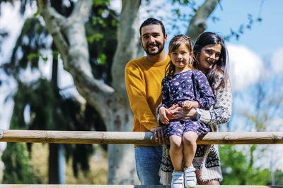 Portrait of smiling parents standing by cute daughter sitting on railing in park