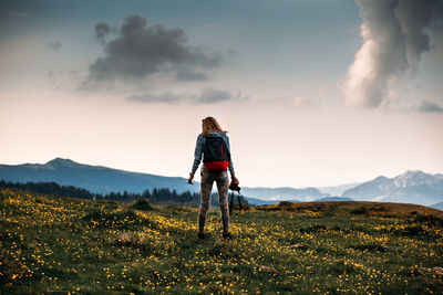 Rear view of woman with backpack standing on landscape against sky during sunset