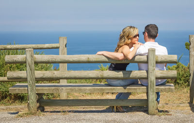 Back view of beautiful love couple sitting outdoors on a bench looking the sea and sky in background