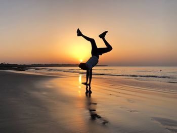 Full length of man doing handstand at beach against sky during sunset