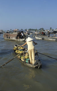 Rear view of female fruit vendor oaring boat in river against clear sky