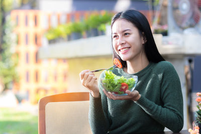 Smiling young woman holding salad bowl while sitting at table