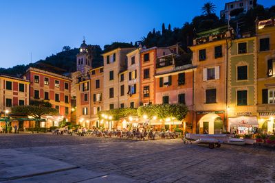 People in illuminated cafe outside buildings at dusk