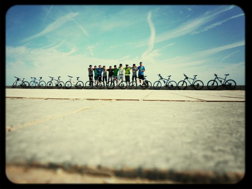 sky, transfer print, men, auto post production filter, leisure activity, sunlight, lifestyles, walking, railing, person, shadow, large group of people, cloud - sky, outdoors, day, cloud, medium group of people, full length, bicycle