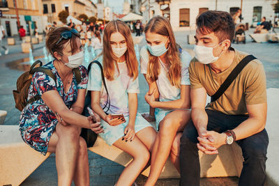 Smiling friends using mobile phone wearing mask sitting outdoors