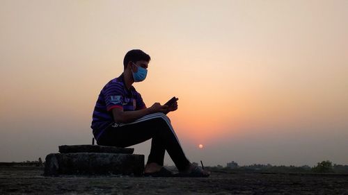 Full length of man wearing mask sitting outdoors during sunset