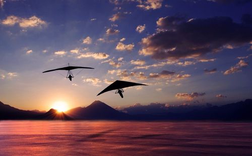 Silhouettes of hang gliders above lake at sunset