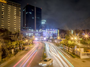 Light trails on city street by buildings against sky at night