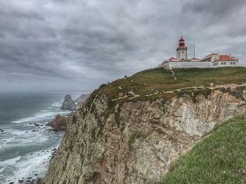 Lighthouse on mountain by sea against cloudy sky