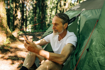 Side view of senior man using mobile phone while sitting in forest